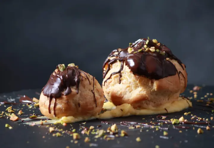 Deux profiteroles au chocolat, garnis de crème pâtissière et saupoudrés de pistaches concassées, sur un fond sombre.