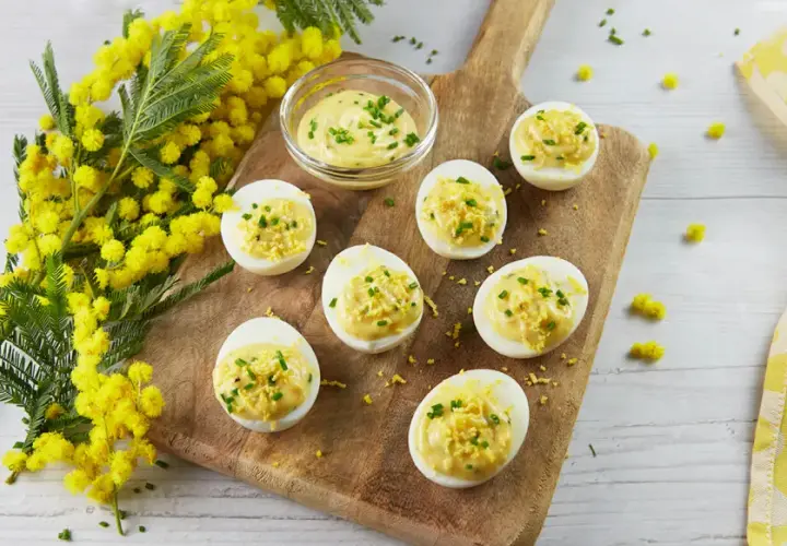 Des œufs mimosa garnis d'une crème onctueuse et de ciboulette, disposés sur une planche en bois, avec des fleurs de mimosa en arrière-plan.