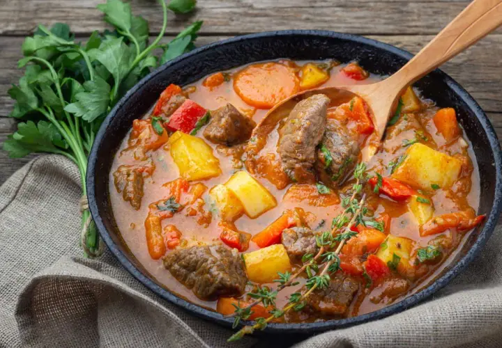 Un plat fumant de recette goulash hongroise traditionnelle, avec des morceaux de bœuf, pommes de terre, carottes et poivrons rouges, servi dans une poêle en fonte avec une cuillère en bois.