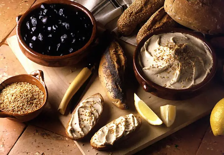 Un bol de purée de pois chiche crémeuse, garnie de graines, avec du pain, des olives noires et des quartiers de citron.