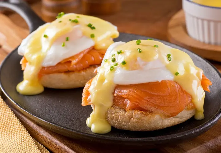 Deux oeufs Bénédicte au saumon fumé, nappés de sauce hollandaise et parsemés de ciboulette, servis sur un muffin anglais dans une poêle.
