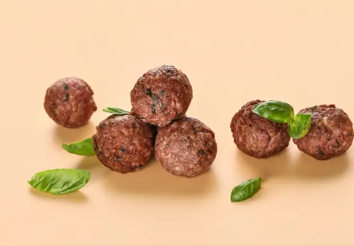 Cinq boulettes de viande dorées, garnies de feuilles de basilic frais, sur fond beige.