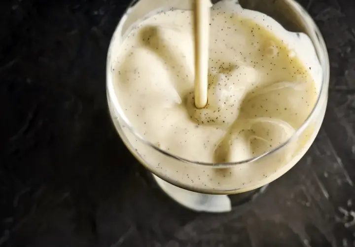 Une crème anglaise onctueuse, parsemée de points de vanille, est versée dans un verre.