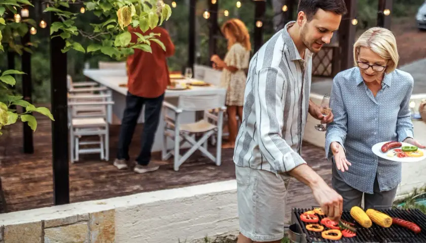 Un homme et une femme préparent un barbecue. Des poivrons, du maïs et des saucisses cuisent sur le gril. Un verre de vin blanc est à portée de main.