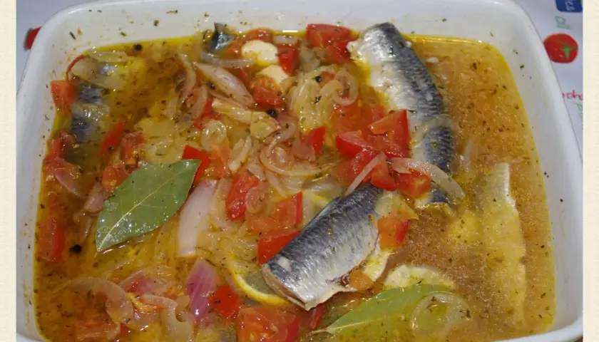 Sardines à l'escabèche dans un plat blanc, avec des tomates, oignons, citron et feuilles de laurier.