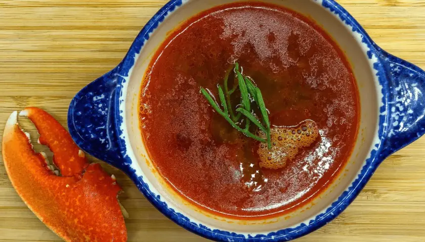 Une délicieuse bisque de homard, garnie d'herbes fraîches, est servie dans un bol bleu et blanc, accompagnée d'une pince de homard sur une planche en bois.