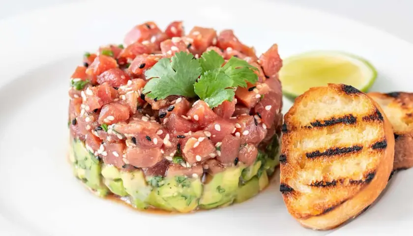 Délicieux tartare de bar frais, garni de sésame et de coriandre, servi avec des toasts grillés et une tranche de citron vert.