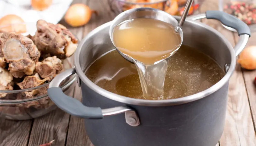 Une louche verse un bouillon doré dans une casserole, sur un fond brun. Des queues de bœuf et des oignons sont disposés autour.