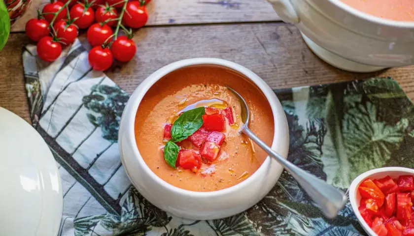 Un bol de soupe à la tomate fraîchement préparée, garnie de dés de tomates et de basilic. Des tomates cerises fraîches sont disposées à côté.