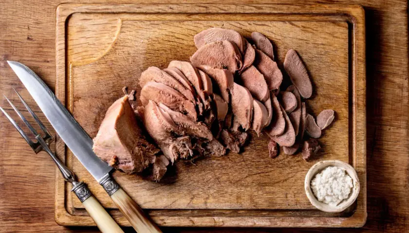 Tranché, la langue de bœuf attend d'être dégustée avec une sauce piquante et des cornichons. Un couteau et une fourchette sont prêts.