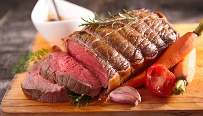 Un délicieux roti de boeuf au four, saignant et tendre, servi avec des carottes, une tomate et de l'ail sur une planche en bois.