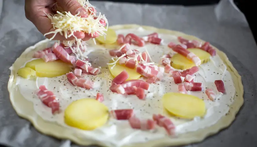 Une main saupoudre du fromage râpé sur une pizza base crème garnie de lardons et de fines rondelles de pommes de terre.