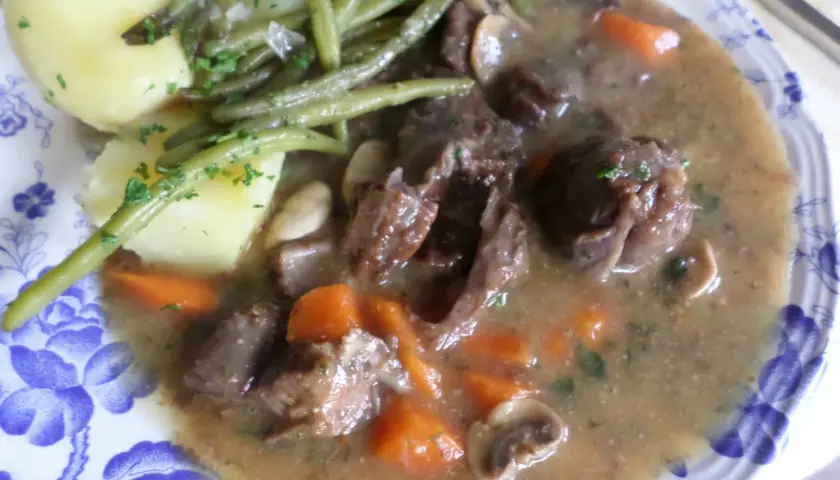 Un plat de civet de daim, avec des morceaux de viande tendres, des carottes, des champignons et des haricots verts, accompagné de pommes de terre.