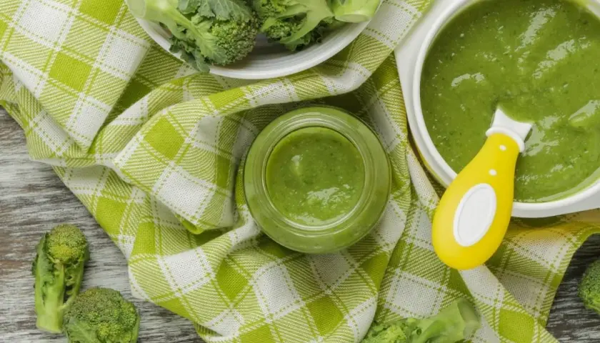 Un bol et un pot en verre remplis de purée de brocolis, avec des fleurettes de brocolis fraîches disposées autour. Une cuillère jaune est dans le bol.
