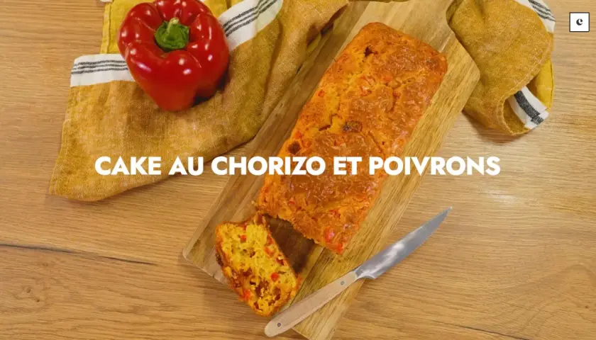 Un cake au chorizo et poivrons, coupé en tranche, sur une planche en bois. Un poivron rouge est posé à côté.