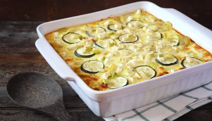 Un plat blanc rectangulaire contient un gratin de courgettes moelleux, garni de rondelles de courgettes et de morceaux de fromage. Une cuillère en bois repose à côté.