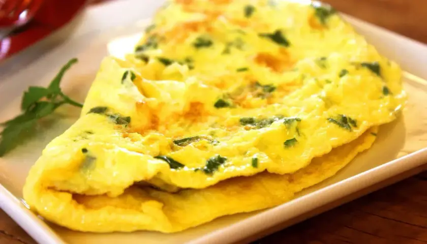 Une omelette au fromage, dorée et moelleuse, parsemée d'herbes fraîches, servie sur une assiette blanche.