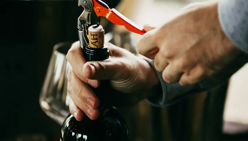 Un homme ouvre une bouteille de vin rouge avec un tire-bouchon. La scène évoque le plaisir de la dégustation et la conservation du vin.