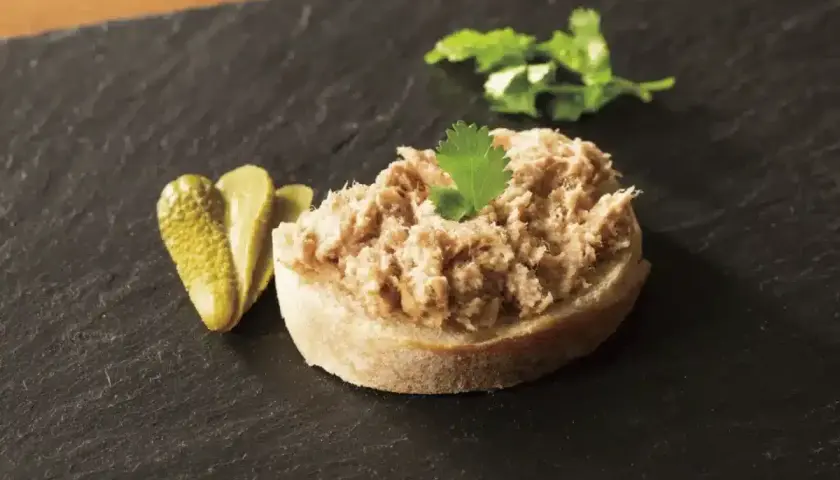 Tranche de pain garnie de rillettes de porc, accompagnée de cornichons et d'une feuille de persil.