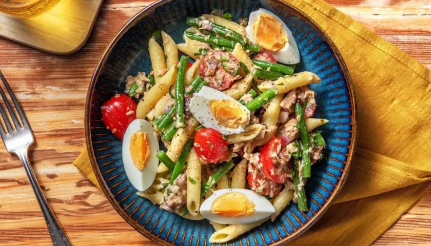 Salade de penne colorée avec haricots verts, tomates cerises, thon et œufs durs, servie dans un plat bleu.