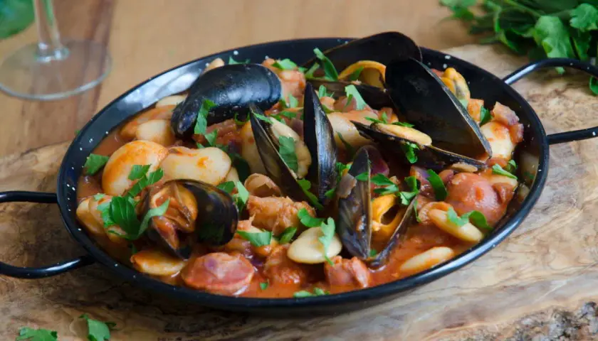 Un plat de moules au chorizo, garni de persil frais, servi dans une poêle noire sur une planche en bois.
