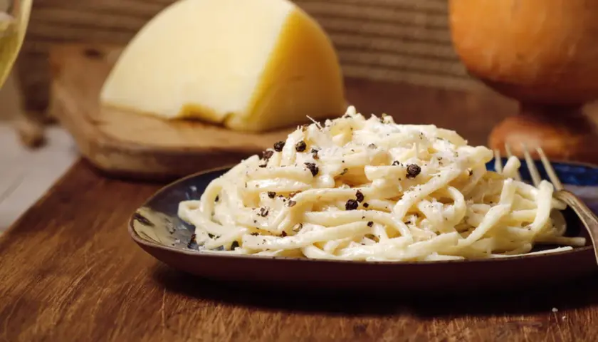Un plat de pâtes crémeux, garni de poivre noir concassé, évoque le goût authentique du cacio e pepe. Un morceau de fromage pecorino est visible en arrière-plan.