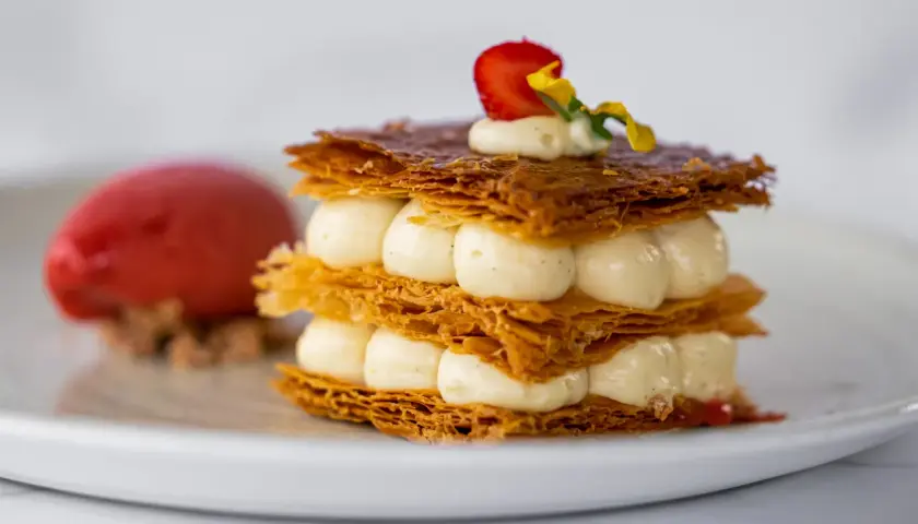 Un délicieux millefeuille croustillant, garni de crème onctueuse et d'une fraise, servi avec une boule de sorbet rouge.