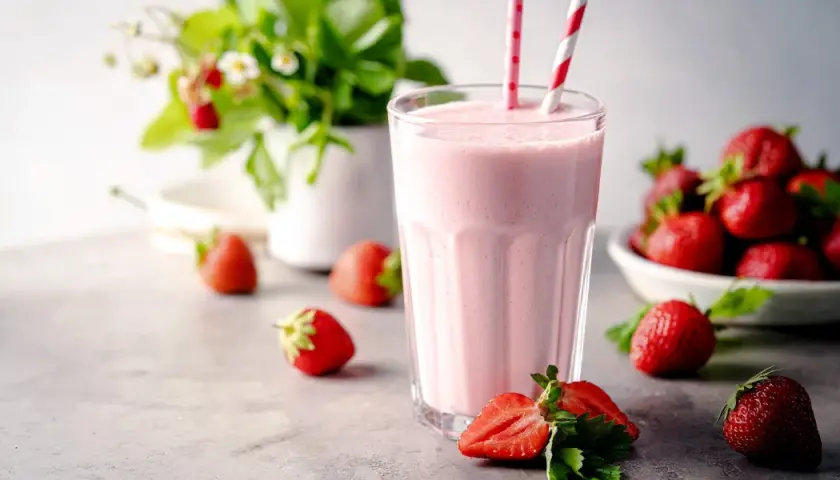 Un délicieux milkshake fraise crémeux, garni de pailles rayées, entouré de fraises fraîches et d'un plant de fraises en arrière-plan.