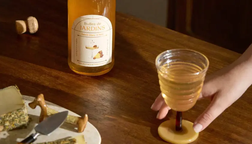 Une main saisit un verre de ce qui pourrait être le meilleur vin blanc moelleux, à côté d'une bouteille "Bulles de Jardins" et de fromages.