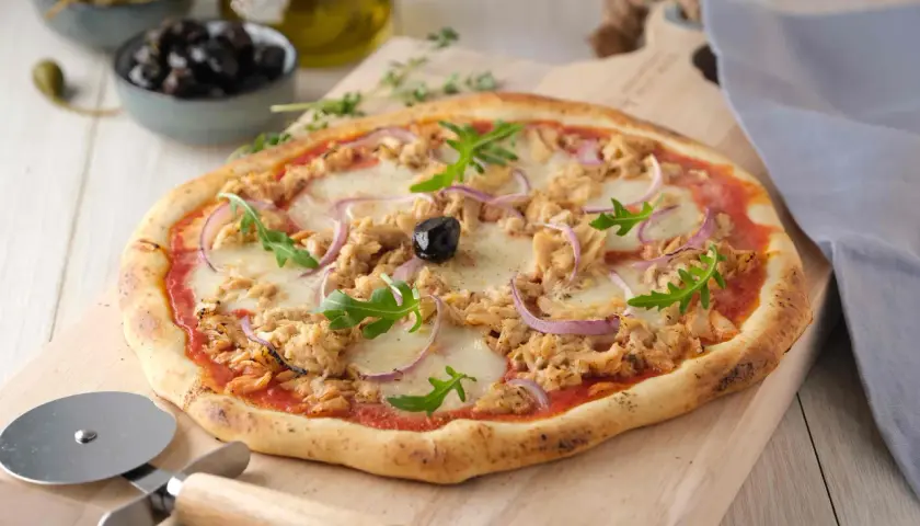 Une délicieuse pizza au thon, garnie d'oignons rouges, de roquette et d'une olive noire, prête à être découpée.