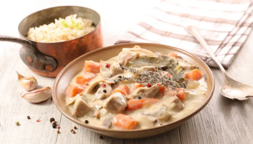 Un plat de blanquette de veau crémeuse avec des carottes et des champignons, servi avec du riz. Un verre de vin rouge est suggéré pour accompagner ce délice.