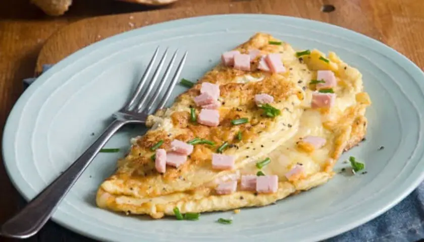Une délicieuse omelette jambon fromage, garnie de ciboulette et de poivre noir, prête à être dégustée avec une tranche de pain croustillant.
