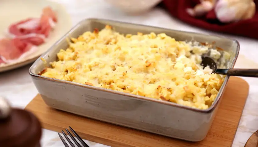 Un plat de gratin de crozet doré et gratiné, prêt à être dégusté. Une assiette de jambon cru est visible à gauche.