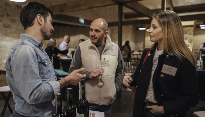 Trois personnes discutent autour d'une table de dégustation. L'une d'elles tient un verre de vin, peut-être un rosé de la terre du midi.
