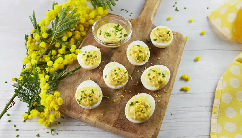 Des œufs mimosa garnis d'une crème onctueuse et de ciboulette, disposés sur une planche en bois, avec des fleurs de mimosa en arrière-plan.