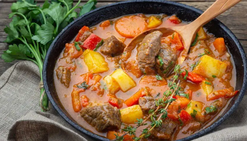 Un plat fumant de recette goulash hongroise traditionnelle, avec des morceaux de bœuf, pommes de terre, carottes et poivrons rouges, servi dans une poêle en fonte avec une cuillère en bois.