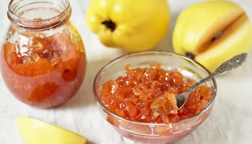 Confiture de coings sans les peler, servie dans un bocal et un bol, avec des coings entiers et une tranche en arrière-plan.