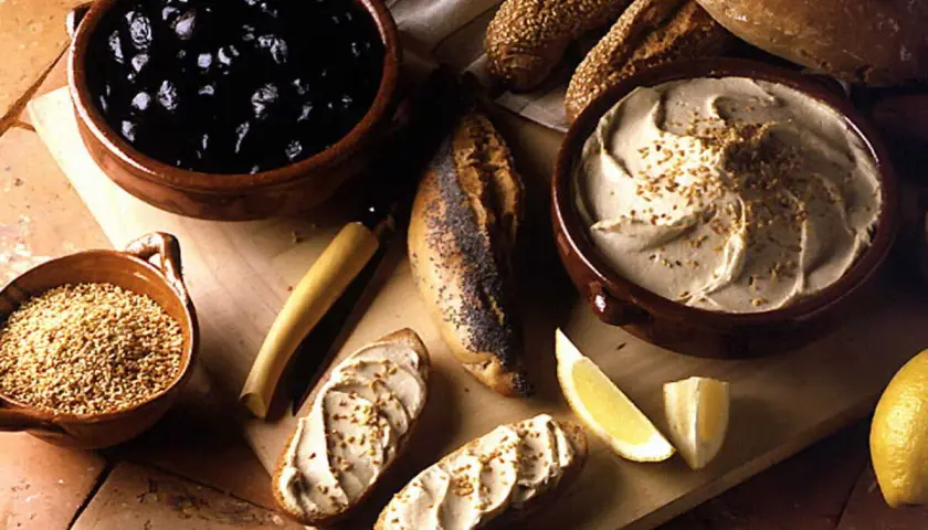 Un bol de purée de pois chiche crémeuse, garnie de graines, avec du pain, des olives noires et des quartiers de citron.