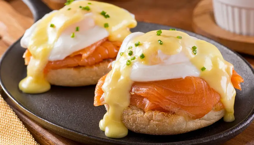 Deux oeufs Bénédicte au saumon fumé, nappés de sauce hollandaise et parsemés de ciboulette, servis sur un muffin anglais dans une poêle.