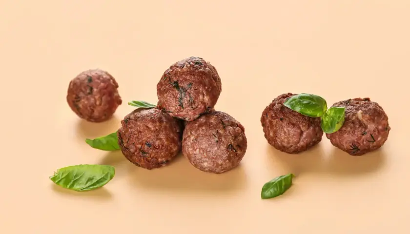 Cinq boulettes de viande dorées, garnies de feuilles de basilic frais, sur fond beige.