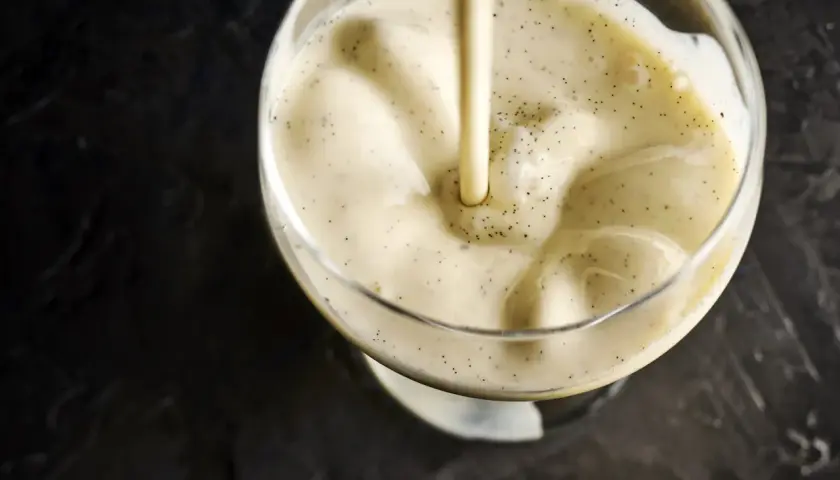 Une crème anglaise onctueuse, parsemée de points de vanille, est versée dans un verre.