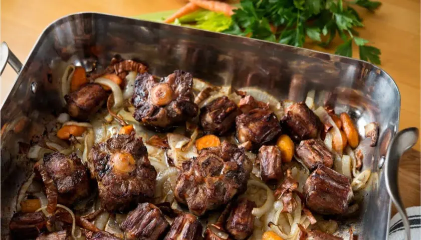 Morceaux de queue de bœuf rôtis dans un plat avec des oignons et des carottes. Un plat savoureux, parfait pour un fond de veau riche.