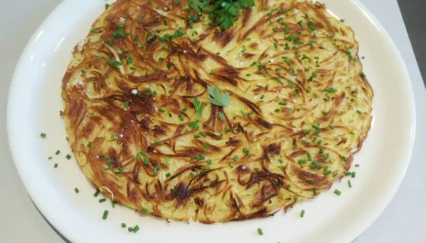 Une pomme darphin dorée et croustillante, garnie de ciboulette fraîche et de persil, servie sur une assiette blanche.