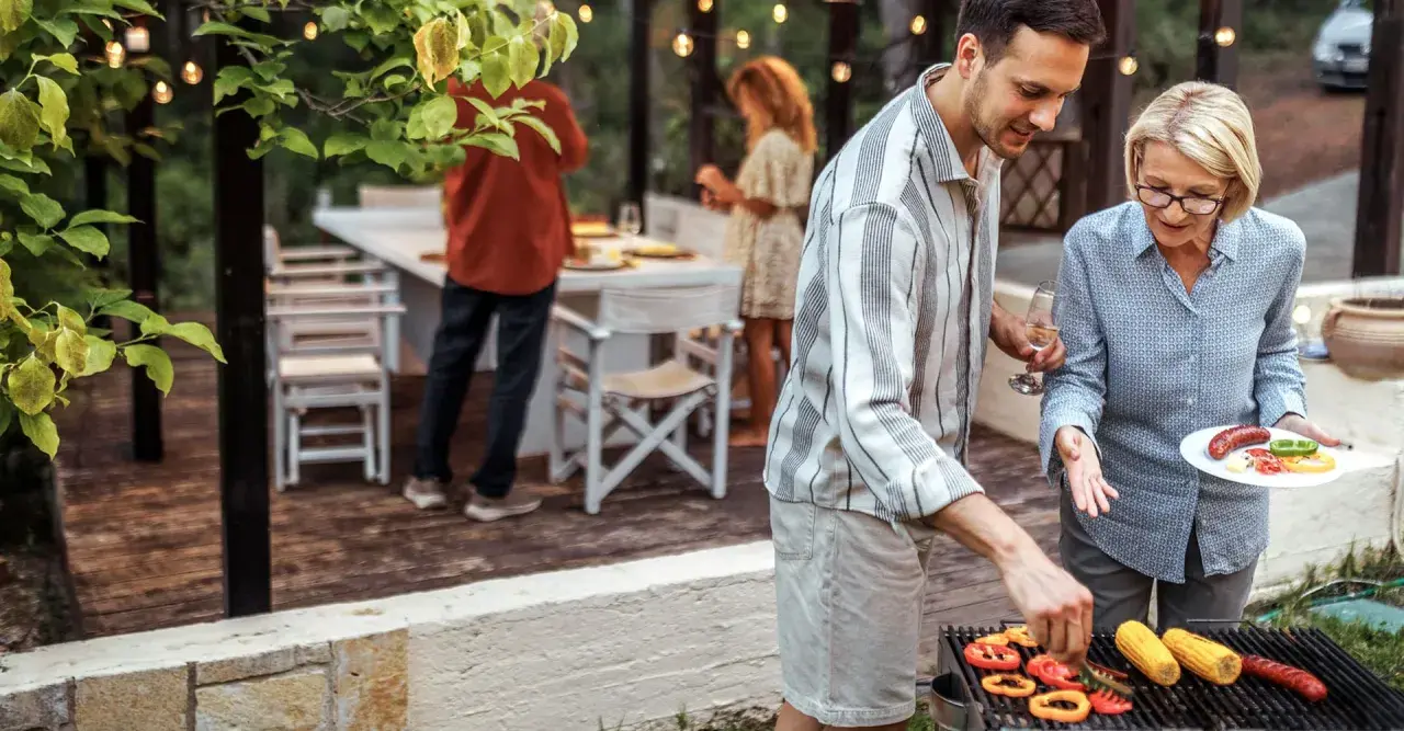 Un homme et une femme préparent un barbecue. Des poivrons, du maïs et des saucisses cuisent sur le gril. Un verre de vin blanc est à portée de main.