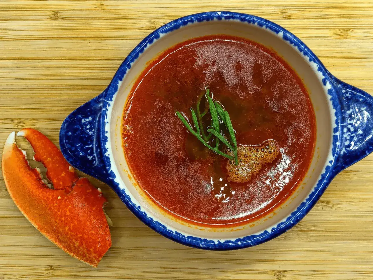 Une délicieuse bisque de homard, garnie d'herbes fraîches, est servie dans un bol bleu et blanc, accompagnée d'une pince de homard sur une planche en bois.