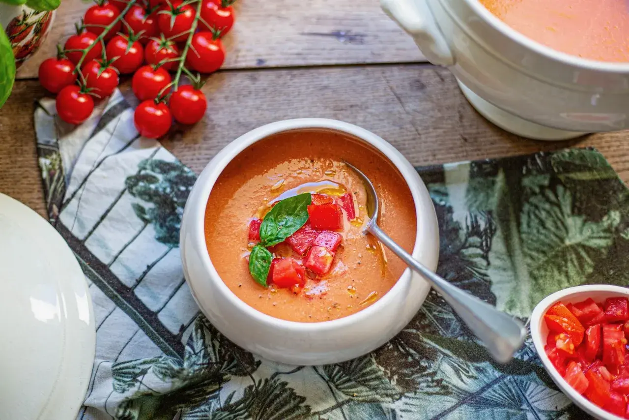 Un bol de soupe à la tomate fraîchement préparée, garnie de dés de tomates et de basilic. Des tomates cerises fraîches sont disposées à côté.