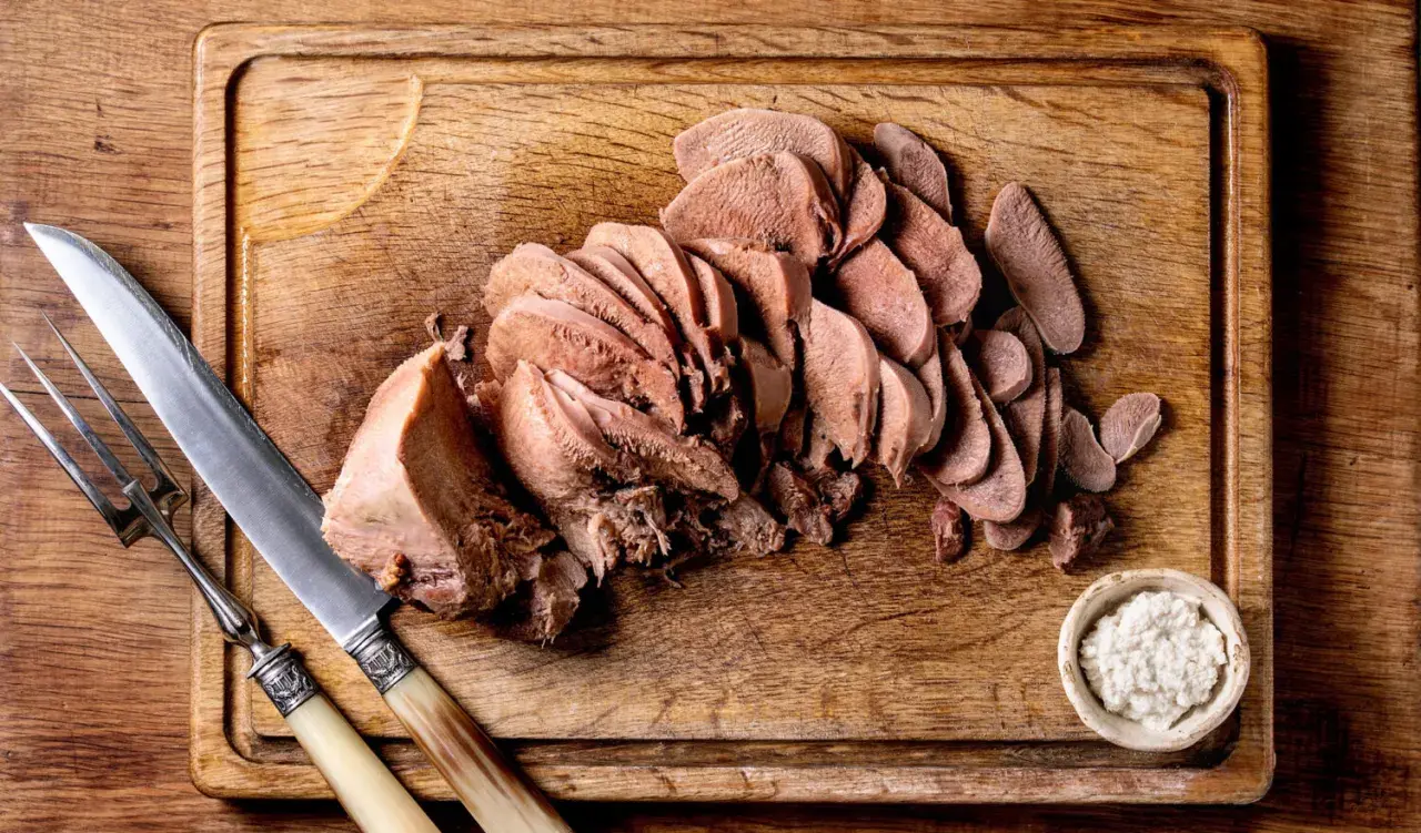 Tranché, la langue de bœuf attend d'être dégustée avec une sauce piquante et des cornichons. Un couteau et une fourchette sont prêts.