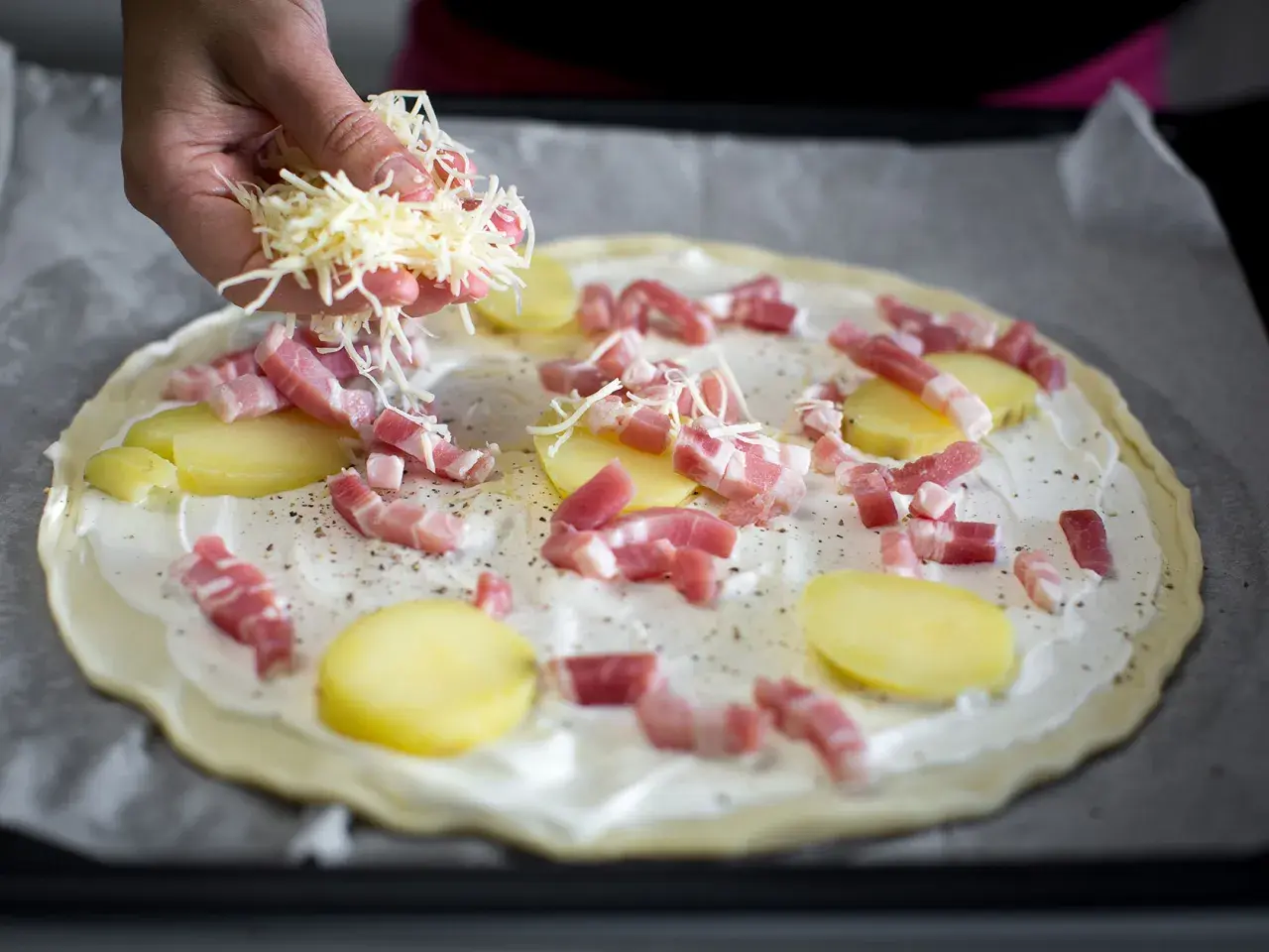 Une main saupoudre du fromage râpé sur une pizza base crème garnie de lardons et de fines rondelles de pommes de terre.