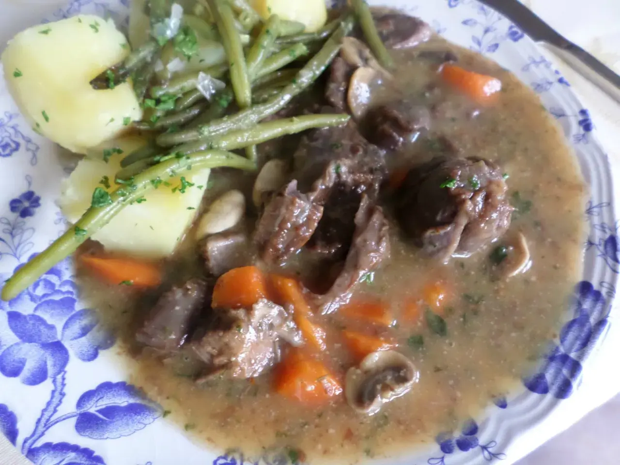 Un plat de civet de daim, avec des morceaux de viande tendres, des carottes, des champignons et des haricots verts, accompagné de pommes de terre.