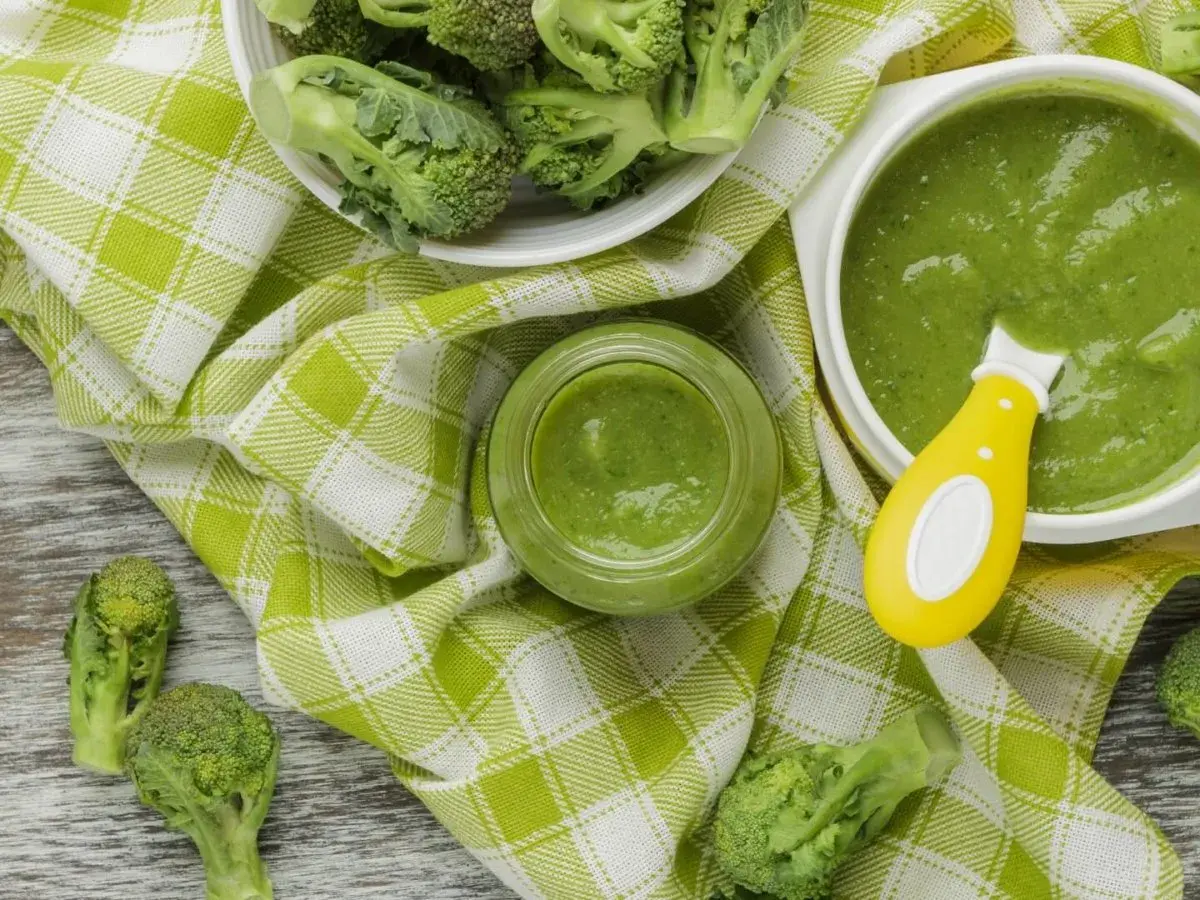 Un bol et un pot en verre remplis de purée de brocolis, avec des fleurettes de brocolis fraîches disposées autour. Une cuillère jaune est dans le bol.